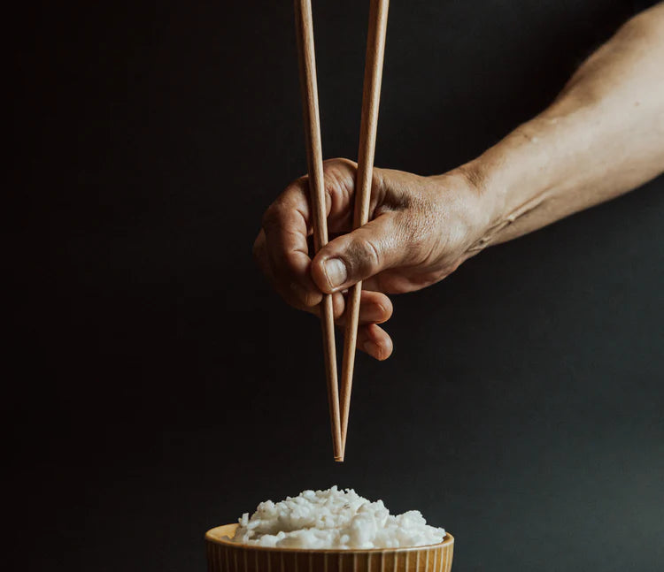 A male-presenting hand holding chopsticks over a bowl of white rice.